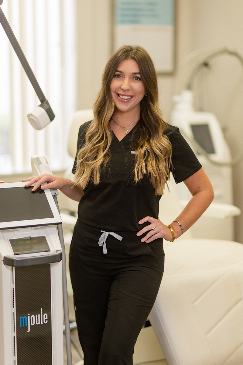 Professional aesthetician in black scrubs posing with a medical-grade laser treatment device at an aesthetic clinic.